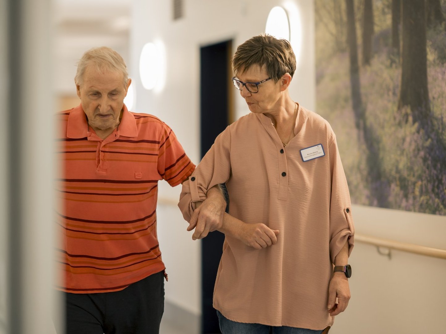 Ein Bewohner spaziert eingehakt mit einer Mitarbeiterin über den Flur.