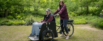 Ein Bewohner fährt gemeinsam mit einer Mitarbeiterin auf einem Rollstuhlfahrrad durch die Natur.