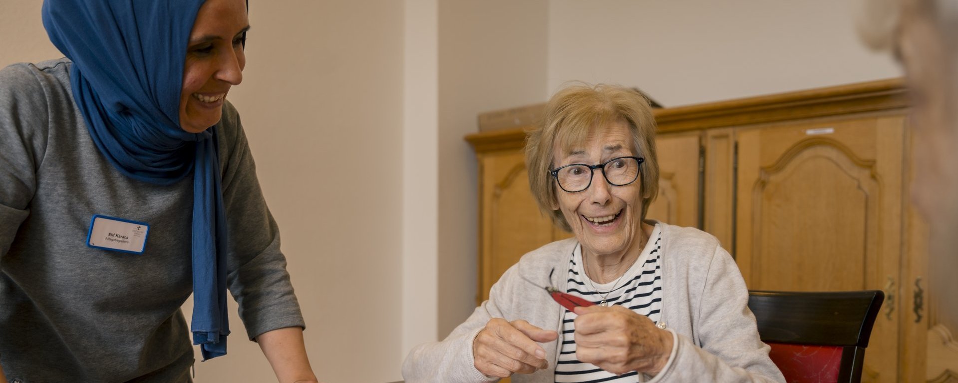 Eine Mitarbeiterin beobachtet lächelnd zwei Bewohnerinnen, die sichtlichen Spaß am Basteln haben.