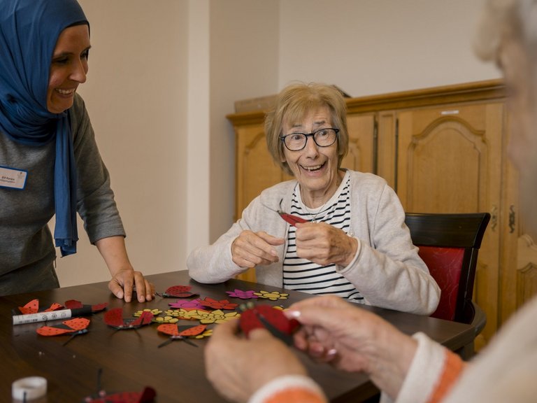 Eine Mitarbeiterin beobachtet lächelnd zwei Bewohnerinnen, die sichtlichen Spaß am Basteln haben.