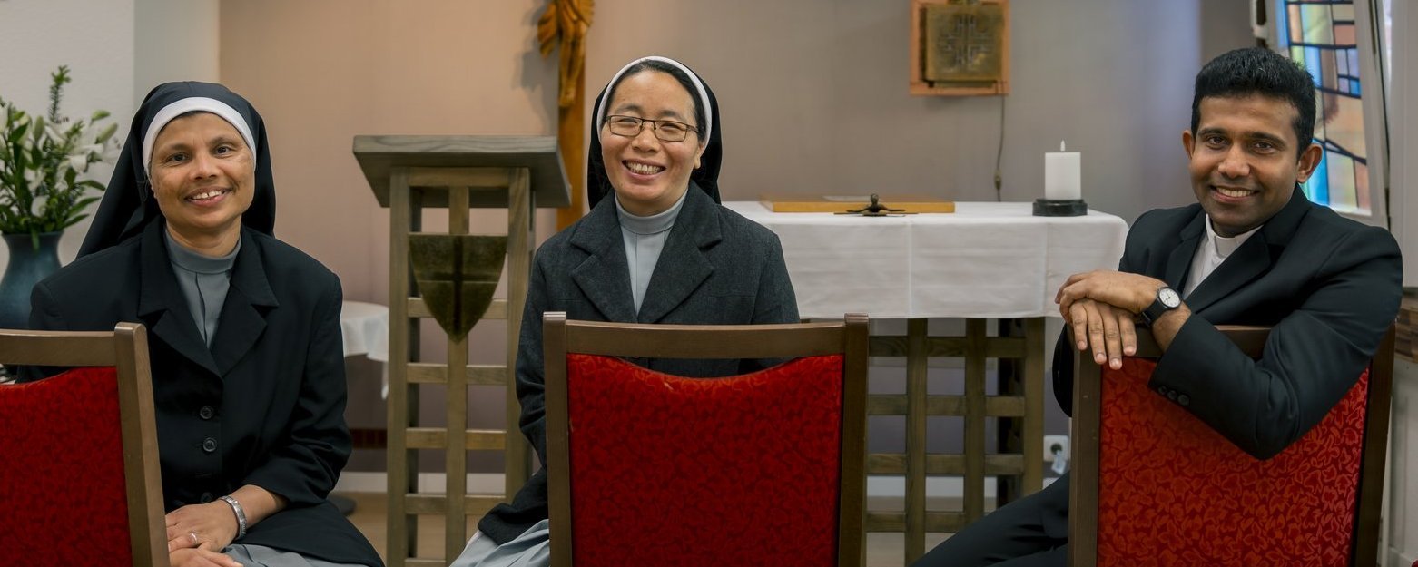 Zwei Schwestern und ein Pater sitzen in der Kapelle und lächeln in die Kamera.