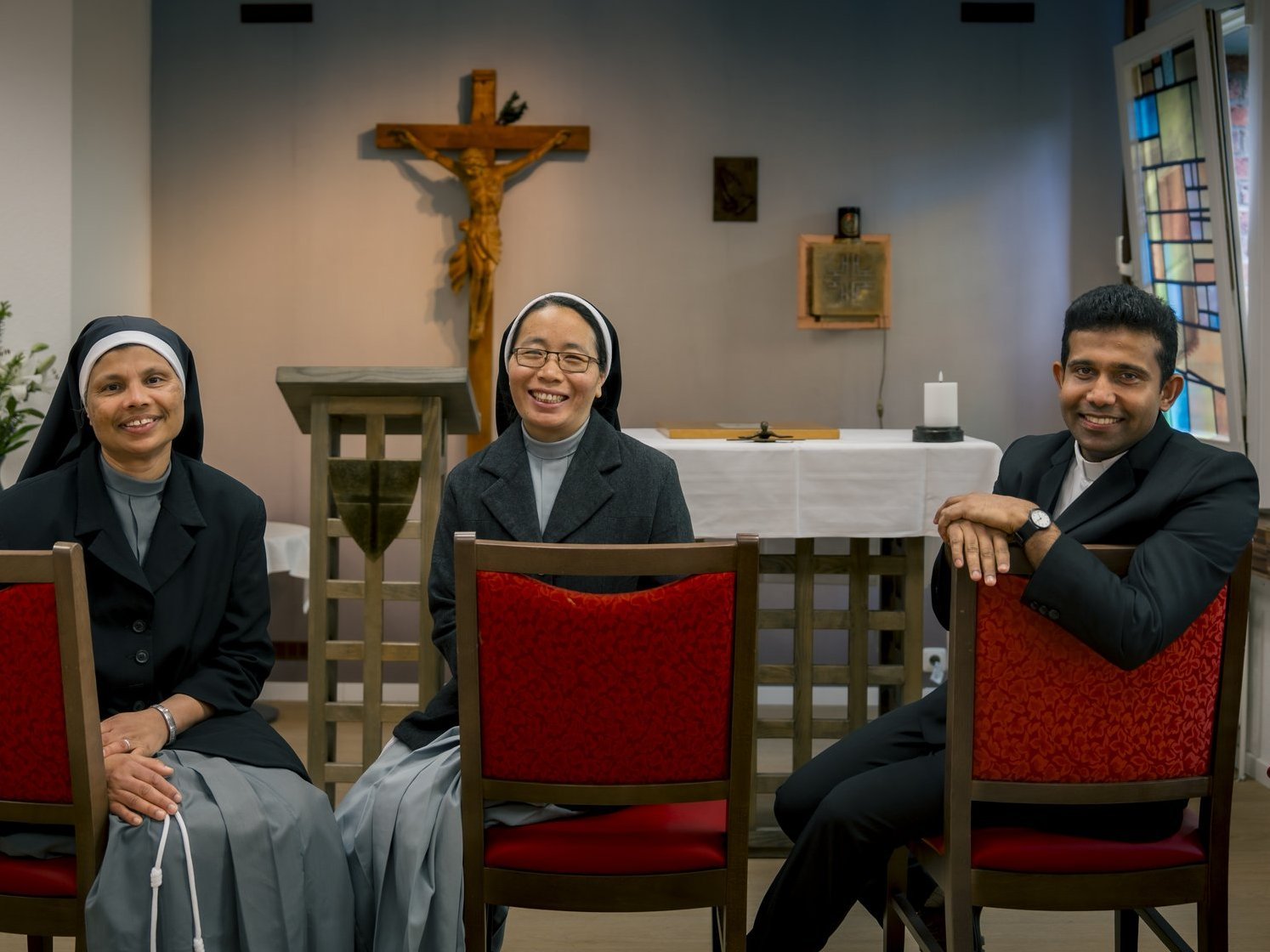 Zwei Schwestern und ein Pater sitzen in der Kapelle und lächeln in die Kamera.