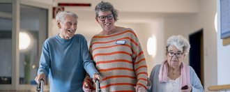 Zwei Bewohnende spazieren mit einer Mitarbeiterin über den Flur. Sie halten sich an den Händen.