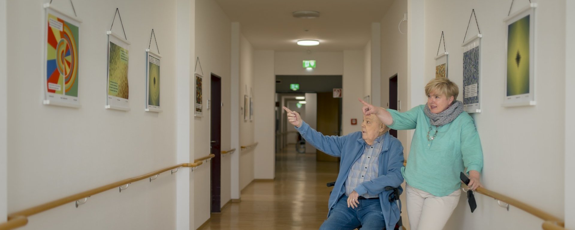 Ein Bewohner betrachtet gemeinsam mit der Quartiersmanagerin seine eigens gefertigten Kunstwerke, die an der Wand hängen.