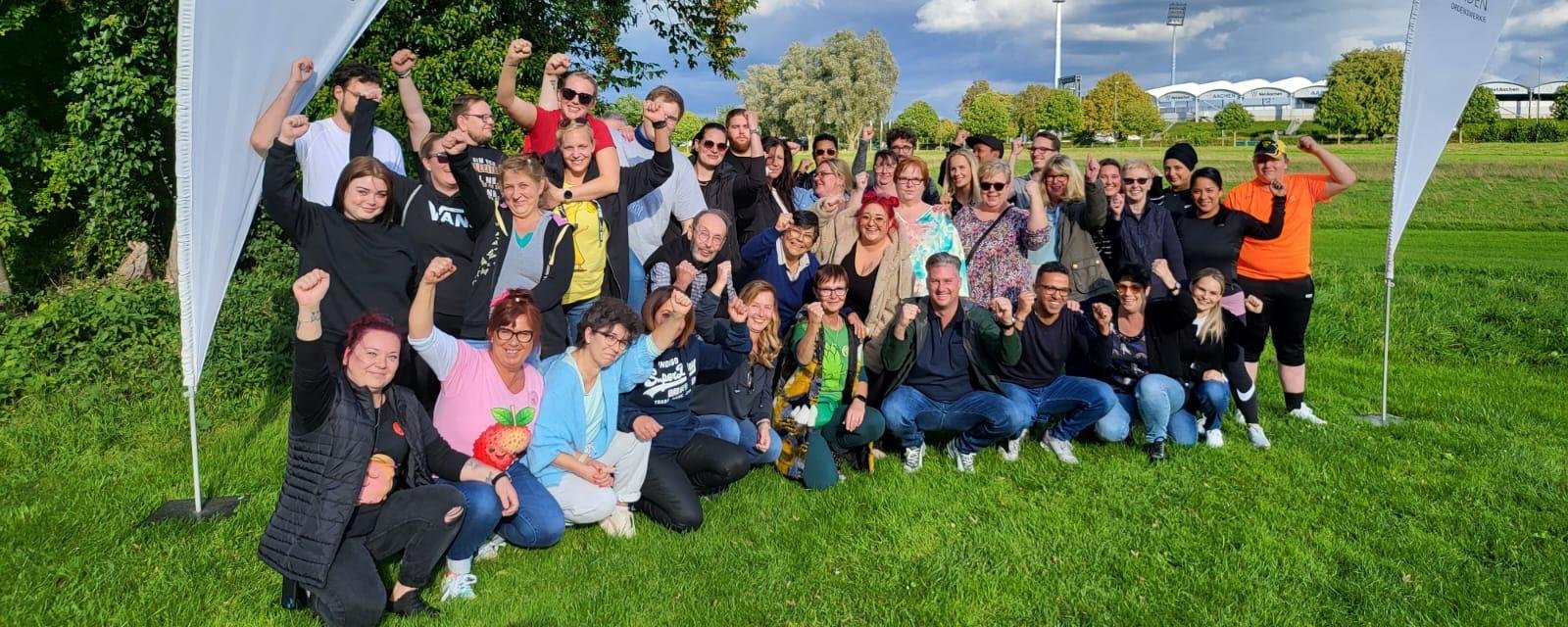 Das bunt gemischte Team des Altenheims Aachen posiert auf einer gruenen Wiese und blauen Himmel für ein Gruppenfoto, umrahmt von Beachflags mit dem St. Raphael Schriftzug zzgl. Logo