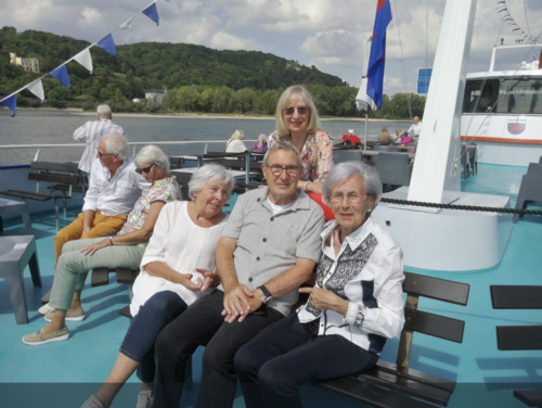 Vier ältere Menschen sitzen auf einem Schiff, im Hintergrund sieht man den Rhein und das gegenüberliegende Ufer mit einem bewaldeten Hügel.